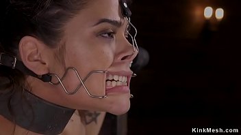 Dark-haired hotty in standing restrain bondage position with wrists and neck in metal stock gets ball-gagged by master